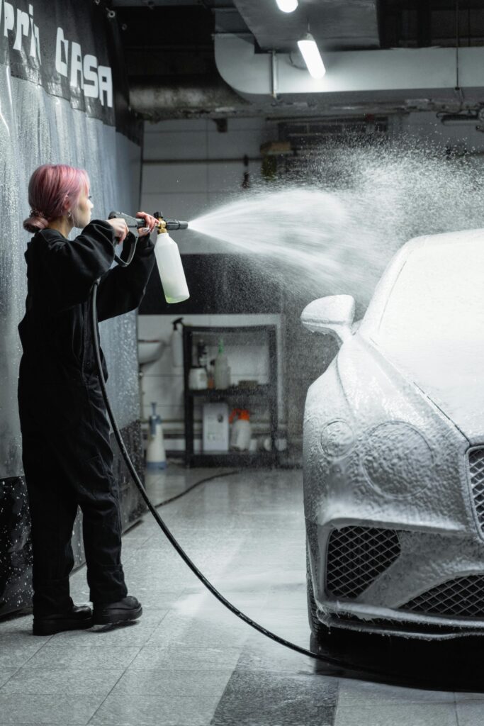 Young woman using a pressure hose to spray foam on a car in a garage setting.